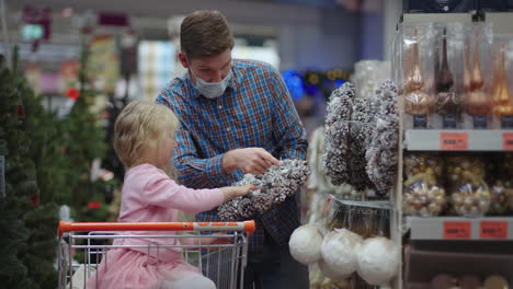 The-girl-sits-in-the-shopping-basket-chooses-a-wreath-with-her-father.-A-father-in-a-medical-mask-on-his-face-with-his-daughter-choose-jewelry-and-toys-on-Christmas-Eve-to-decorate-the-house